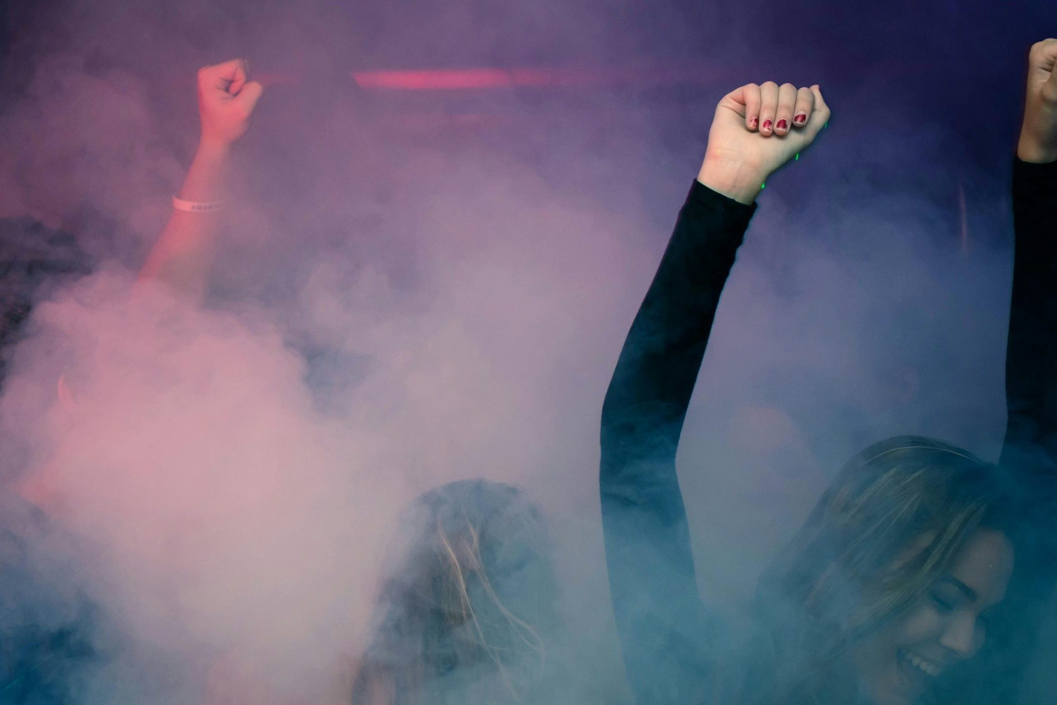 Crowd of people dancing energetically in a foggy nightclub with raised hands.